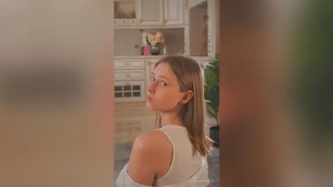 Young woman in a white sleeveless top looking thoughtfully in a bright, modern kitchen setting.