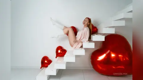 Cheerful woman in pink loungewear sitting on white stairs surrounded by red heart balloons.