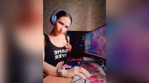 Young woman in black graphic tank top wearing blue headphones while gaming at a desk setup.