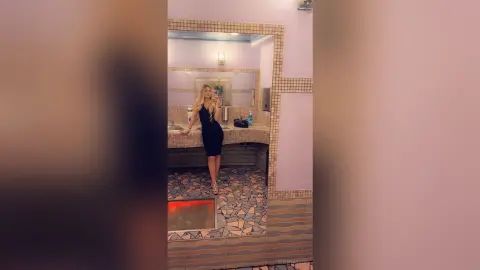 Blonde woman in a chic black cocktail dress taking a mirror selfie in a modern tiled restroom.
