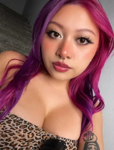 Young woman with vibrant purple hair and winged eyeliner wearing a leopard print top in a soft indoor light.
