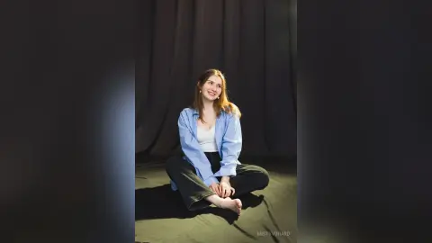 Smiling young woman in a blue striped shirt sitting on a dark floor in a moody studio setting.