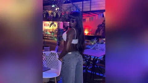 Stylish Black woman in a white bikini top and denim jeans posing at a vibrant night rooftop lounge.