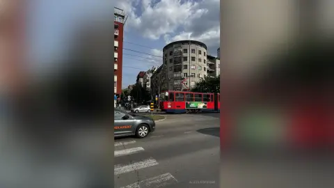 Red city tram driving past modern apartment buildings on a bright urban street during the day.