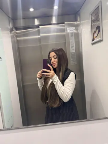 Young woman with long hair taking a selfie in an elevator wearing a white long-sleeve shirt and navy pinstripe vest.