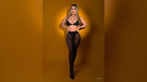 Lucine poses in a black bra and matching black panties against a solid gold background, showing off her long hair and slim figure.
