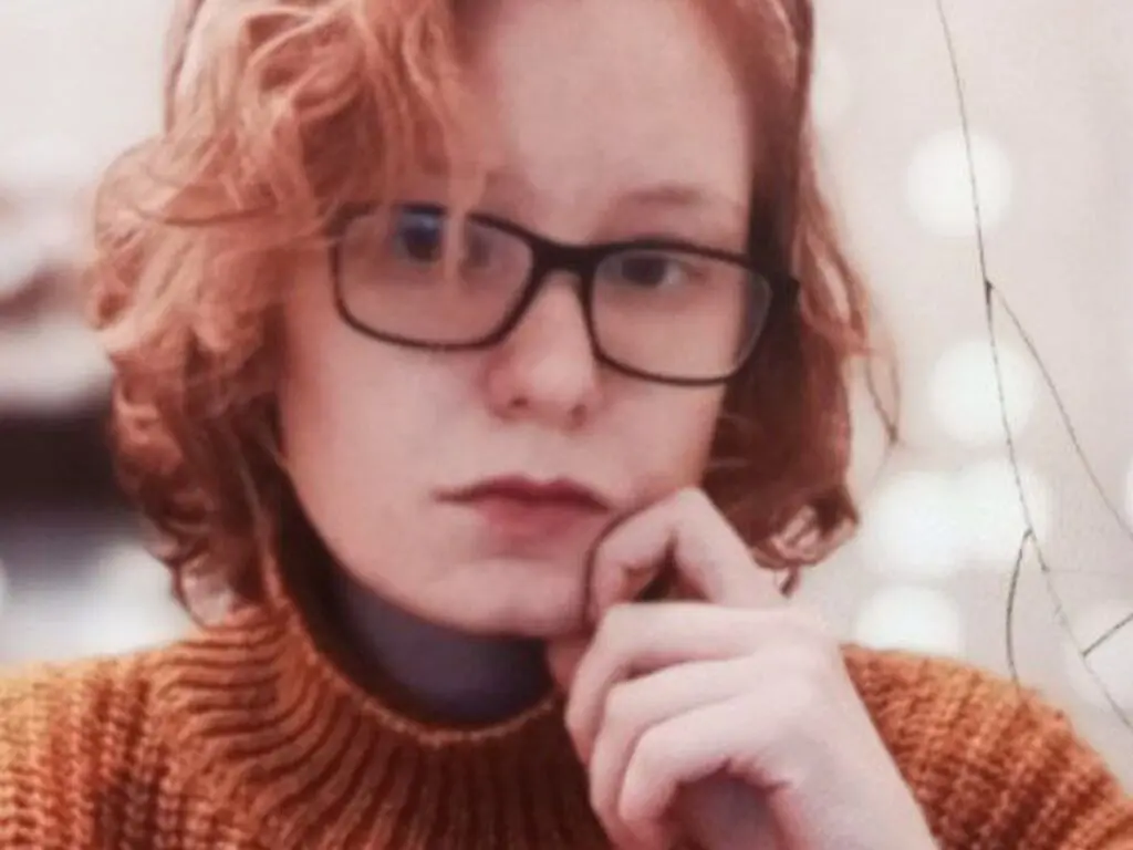 LeaShari wearing black glasses and a brown knit sweater, resting her chin on her hand with her wavy red hair down.