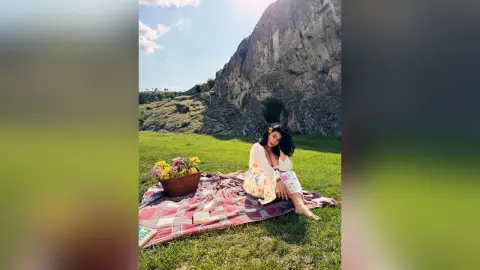 Woman in floral outfit sitting on a picnic blanket with flowers near a large mountain under a blue sky.
