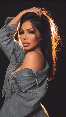 Glamorous woman in a denim jacket with long dark hair posing against a dark studio background.