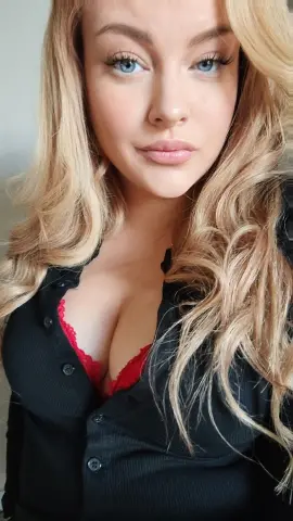 Blonde woman with blue eyes wearing a black shirt and red lace lingerie in a soft indoor setting.