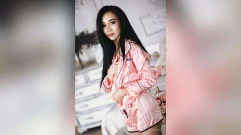 Young woman with long black hair wearing pink striped pajamas in a bright, cozy bedroom setting.