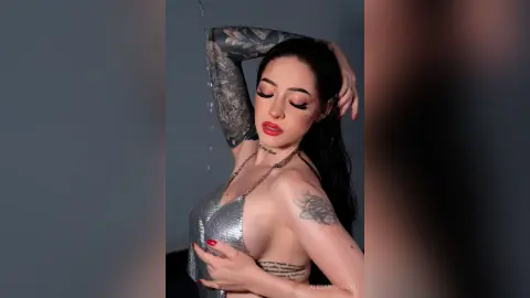 Tattooed woman in a silver sequin top posing under falling water in a moody studio setting.