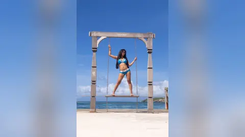 MokaAkashiya — Stylish woman in a blue bikini relaxing on a wooden swing at a sunny tropical beach resort.