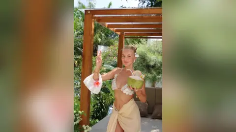 Smiling woman in a cream bikini holding a coconut drink under a tropical wooden pergola.