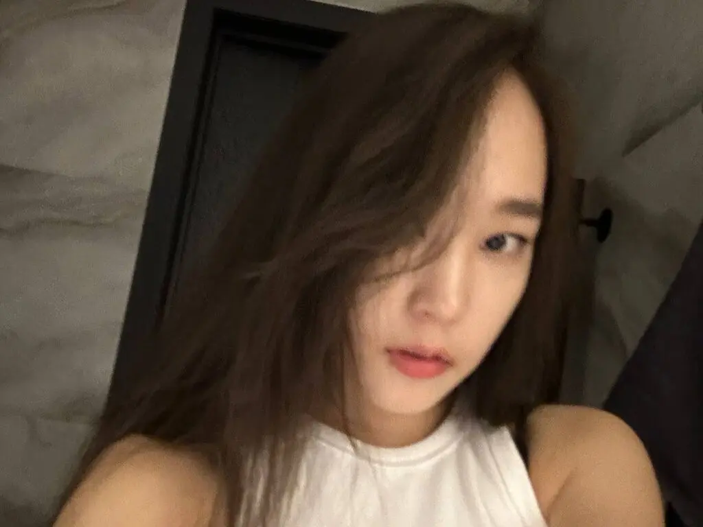 Young woman with long brown hair wearing a white sleeveless top in a moody indoor setting.