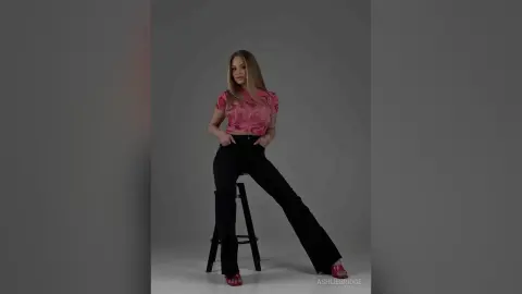 Blonde woman in a pink patterned crop top and black flared jeans sitting on a stool against a grey background.