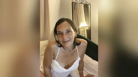 MichaelaBelle smiles while wearing a white lace camisole, leaning forward on a bed with a lamp and mirror in the background.