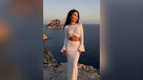 Smiling woman in a white long-sleeve crop top and maxi skirt standing on a rocky cliff overlooking the ocean at sunset.