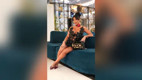 Dolce sits on a blue velvet sofa wearing a black lace cutout bodysuit and red heels.