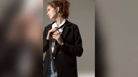 Young woman in a black blazer and white shirt adjusting her tie against a minimalist grey background.