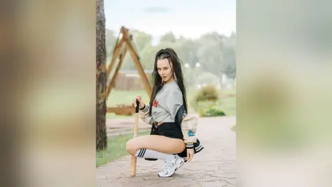 Young woman in casual streetwear posing with a baseball bat in a sunny outdoor park setting.