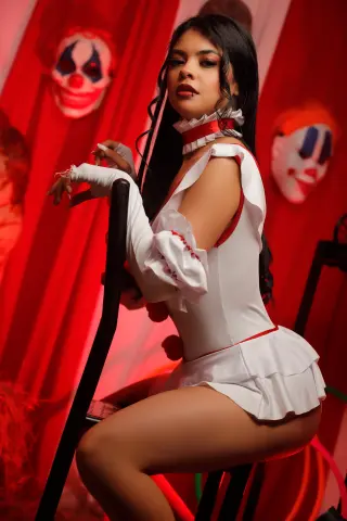 EmilyyJhonson wears a white ruffled clown outfit and sits against a red curtain background with clown masks.