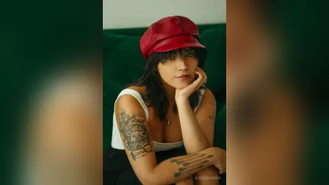 Tattooed woman in a white tank top and red newsboy cap posing thoughtfully against a green velvet background.