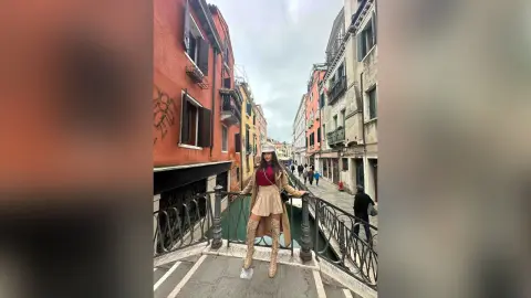 Stylish woman in a beige trench coat and boots walking across a narrow stone bridge in a historic European city.