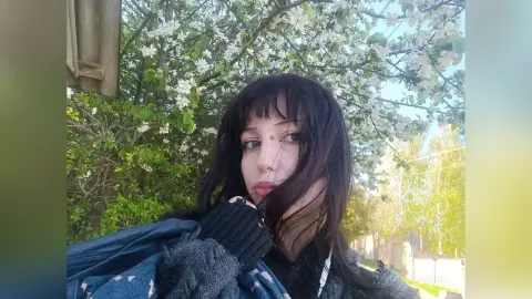 Young woman with dark hair and bangs in a cozy black sweater outdoors under blooming white spring flowers.