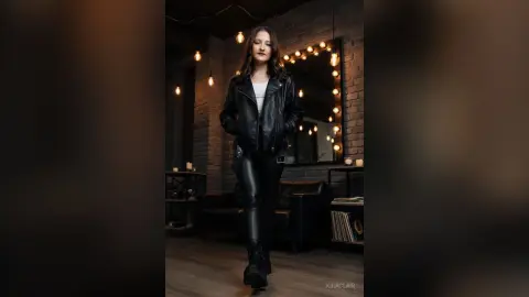 Stylish woman in a black leather jacket and pants posing in a moody, dimly lit industrial loft setting.