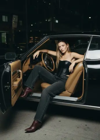 Stylish woman in black corset and jeans sitting in a vintage car at night for a moody urban fashion look.