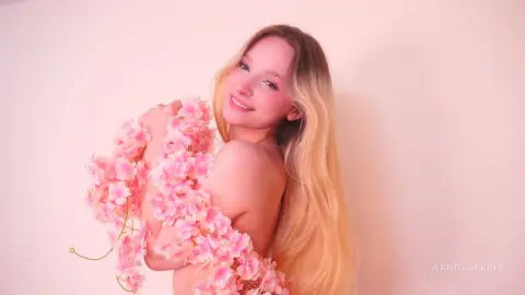 Smiling blonde woman draped in pink artificial flowers against a soft pink background.
