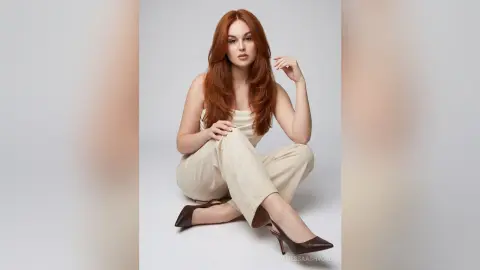 Elegant redhead woman in beige jumpsuit sitting on a white studio floor with a sophisticated mood.