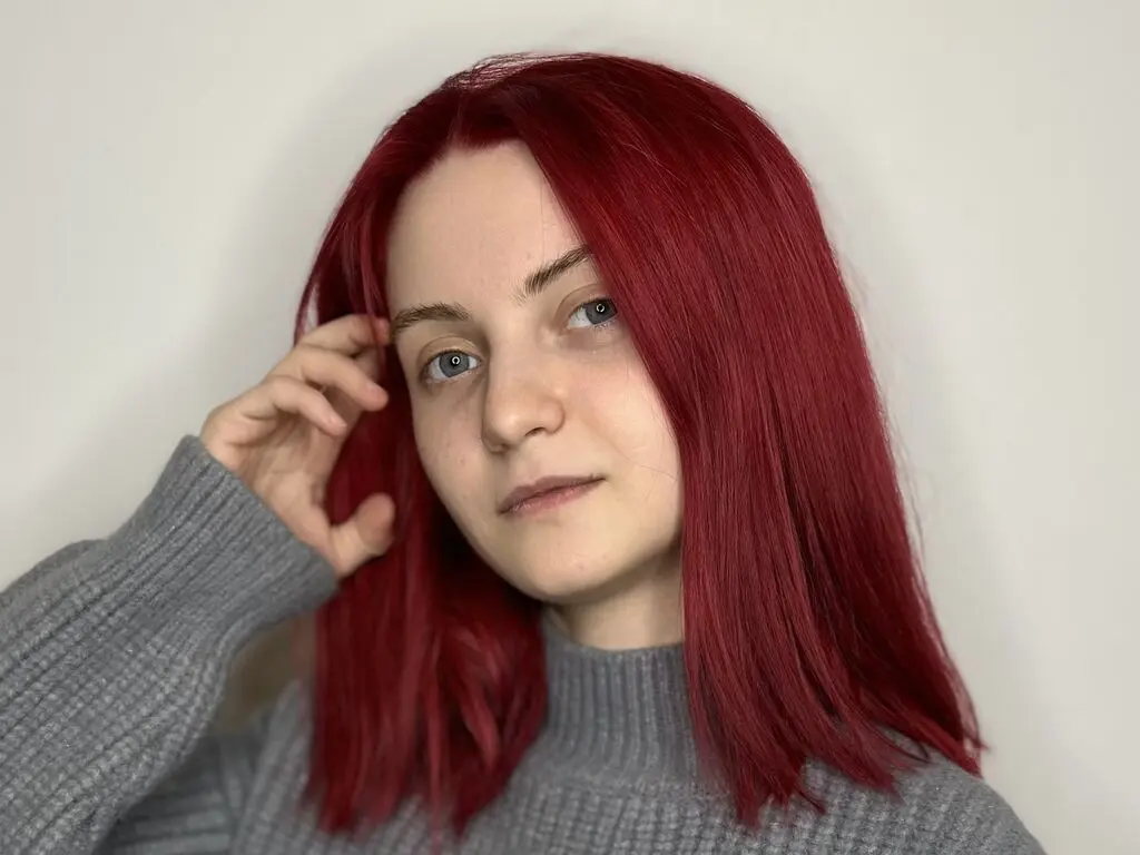 Young woman with long red hair wearing a grey ribbed sweater against a plain white background.