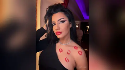 MissyJolie poses with long dark hair and red lipstick, wearing a black top and a silver necklace with red lipstick kisses on her shoulder.