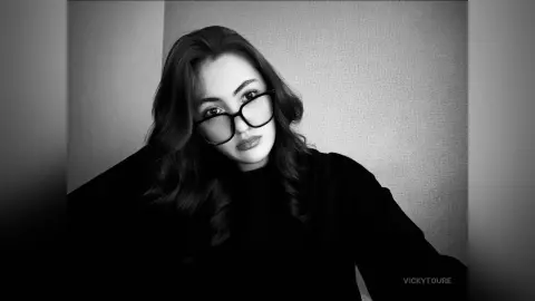 VickyToure poses in a black top and thick black glasses, looking at the camera with long, wavy dark hair against a plain wall.