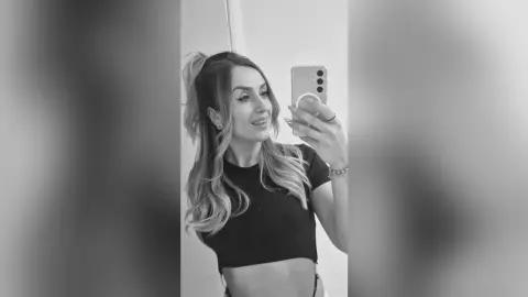 Smiling young woman in a black crop top taking a casual mirror selfie in a bright room.