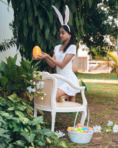 Young woman in a white dress holding a golden Easter egg while sitting outdoors in a lush garden setting.