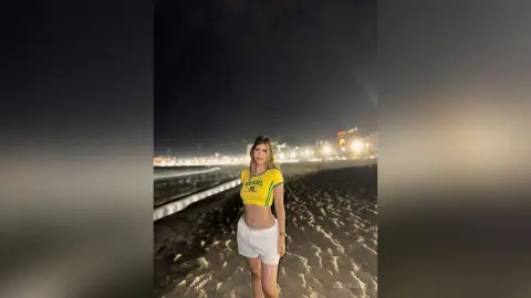 Young woman in a yellow Brazil crop top and white shorts standing on a dark beach at night.