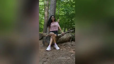 Smiling woman in a pink t-shirt and black shorts sitting on a log in a lush green forest.