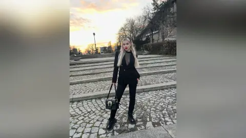 Blonde woman in chic black blazer and trousers posing on stone steps at sunset.