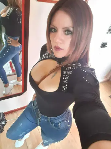 Woman in black studded top and blue jeans taking a selfie in a bright room.