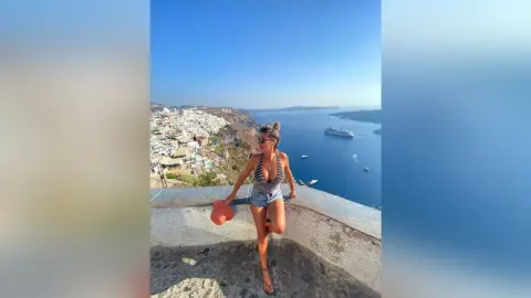 AlexiaMendoza poses outdoors in a low-cut black bodysuit and denim shorts, overlooking a bright blue sea and coastal town.