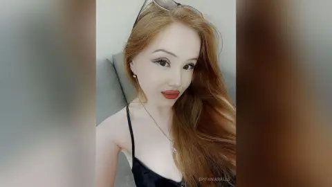EpifaniaRalls poses with long reddish-blonde hair, wearing a black camisole and a silver necklace against a grey sofa background.