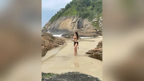 Woman in a black bikini standing on a rocky tropical beach with ocean waves and lush green cliffs.