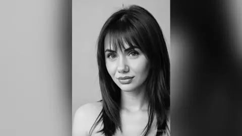 Portrait of a young woman with long dark hair and bangs looking serene in a black and white studio setting.