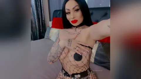 DeeaAnderson poses in black fishnet lingerie and a black choker, showing her chest and tattooed arms while resting on a bed.