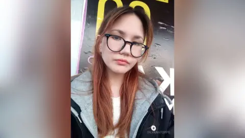 Young woman with long red hair and black glasses wearing a grey hoodie in a casual indoor setting.