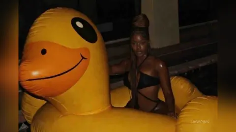 Young woman in black bikini relaxing next to a giant yellow inflatable duck float in a dark poolside setting.