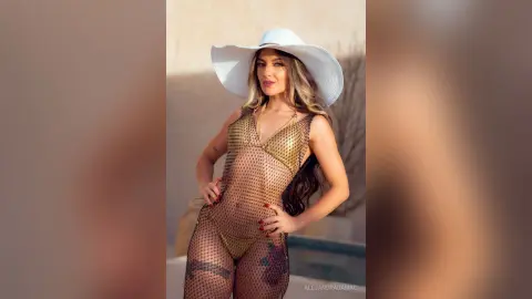 Glamorous woman in a gold mesh bodysuit and wide-brimmed white sun hat posing in bright sunlight.